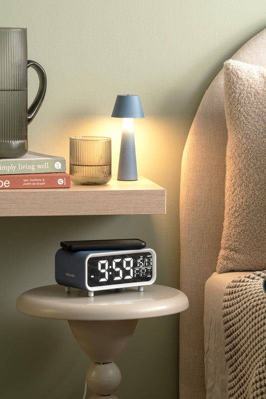 Digital clock on a nightstand next to a bed with a lamp and books in the background.