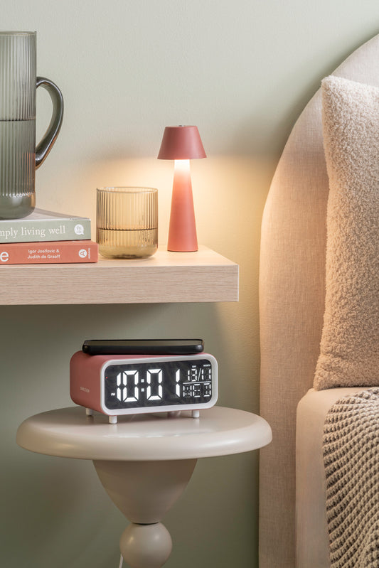 Pink alarm clock on a nightstand with a lamp and books in the background