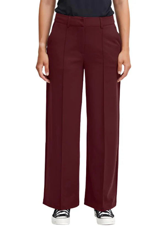 A pair of burgundy long trousers with a crease down the front, flat waistband, and back jetted pockets, worn by a person standing against a white background.