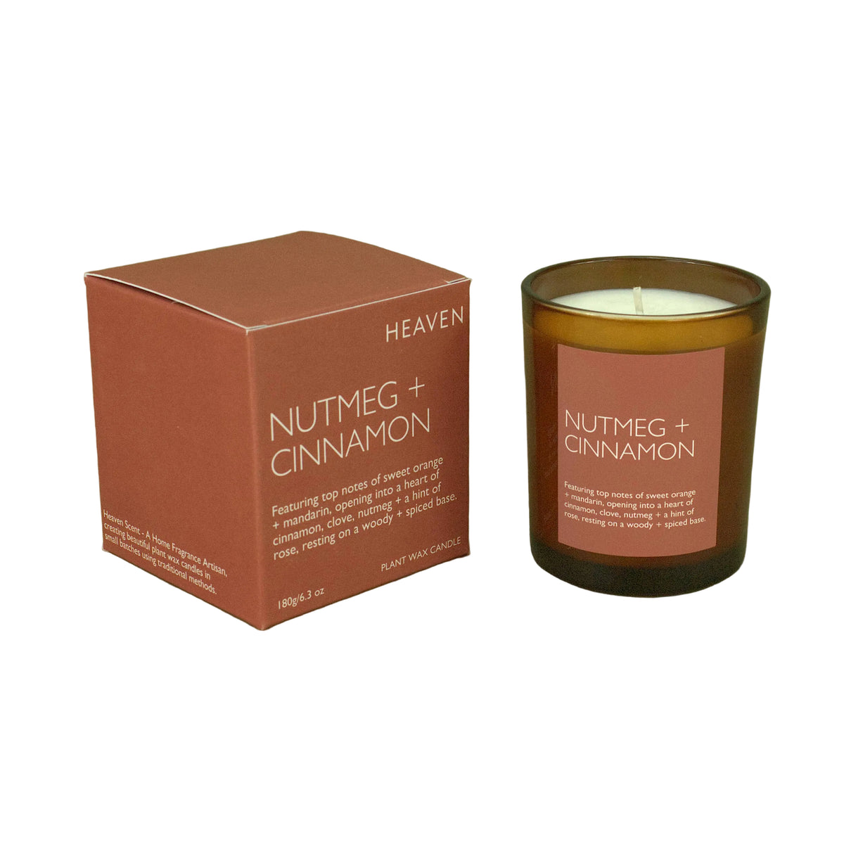 A candle in a brown glass jar with a pink label next to its matching pink box, labeled 'HEAVEN NUTMEG + CINNAMON', set against a solid olive green background.