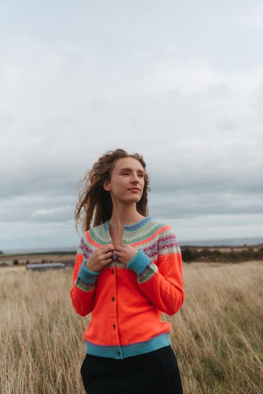 A woman stands in a grassy field under a cloudy sky, wearing a bright orange cardigan with a multicolor Fair Isle pattern at the collar and cuffs, paired with a black skirt.