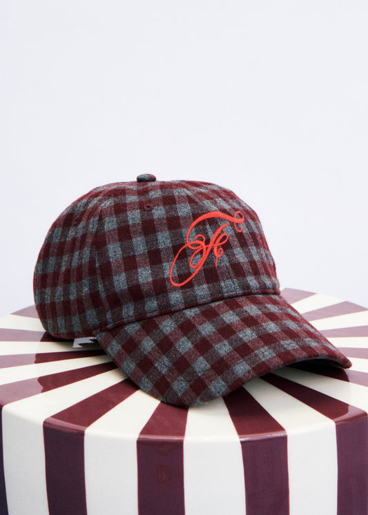 A plaid baseball cap in maroon and gray with a red embroidered monogram logo on the front, displayed on a striped round pedestal against a light gray background.