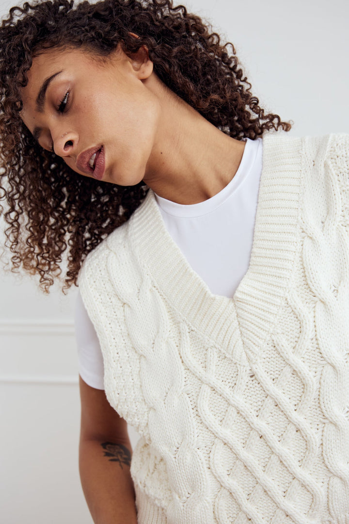 Noella Gio Knit Vest in Ivory. Woman wearing a cream cable knit vest over a white shirt against a white background