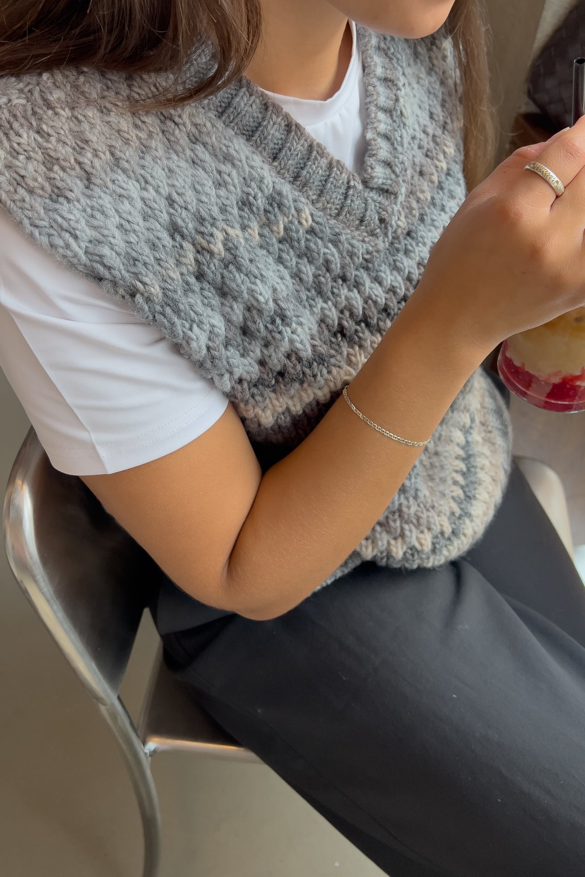 Noella Gio Knit Vest in Light Grey Mix. Person wearing a grey knitted vest sitting on a chair, holding a phone.