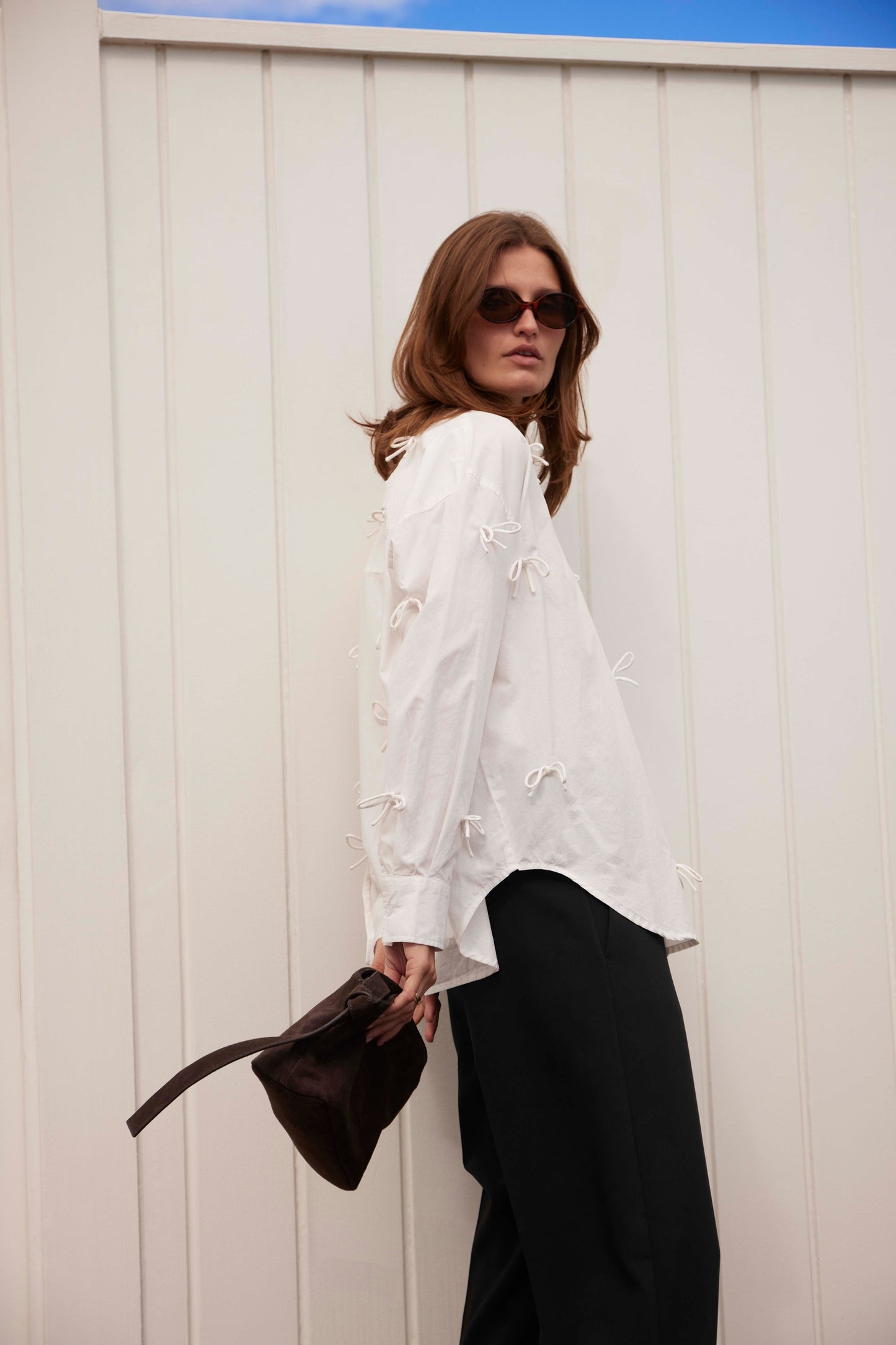 Soaked in Luxury Joa Shirt in Broken White. Woman in a white blouse and black pants holding a brown bag against a white wall.