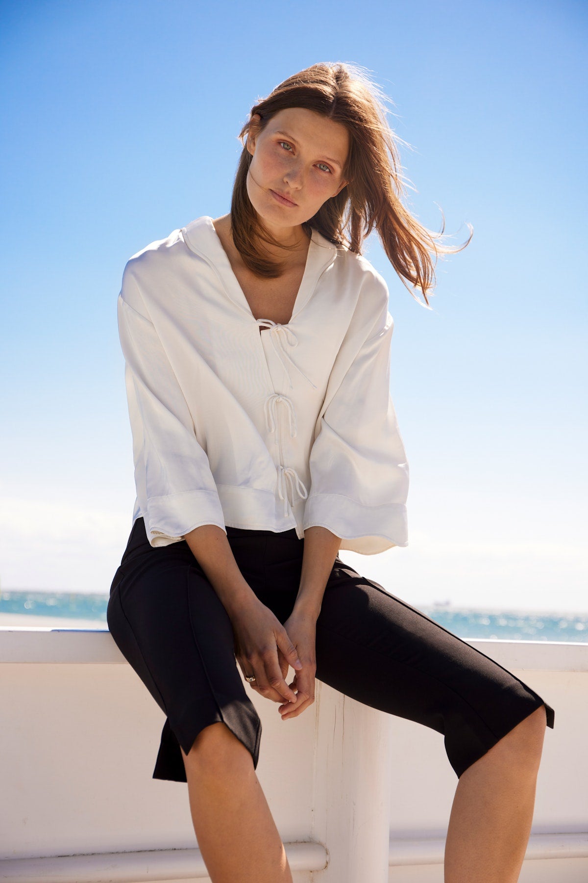 Soaked in Luxury Leena Elia SS Shirt in Whisper White. Woman wearing a white shirt and black shorts sitting on a ledge with a clear blue sky and ocean background.