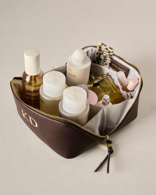 A brown zippered cosmetic bag with gold hardware and a monogrammed 'KD' on the front, shown open and filled with assorted skincare and perfume bottles on a light beige background.