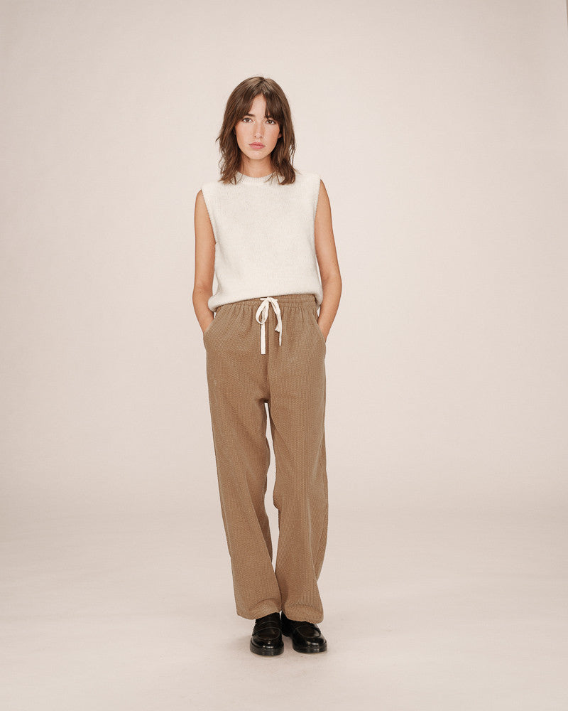 Woman wearing a sleeveless white top and beige pants on a plain background