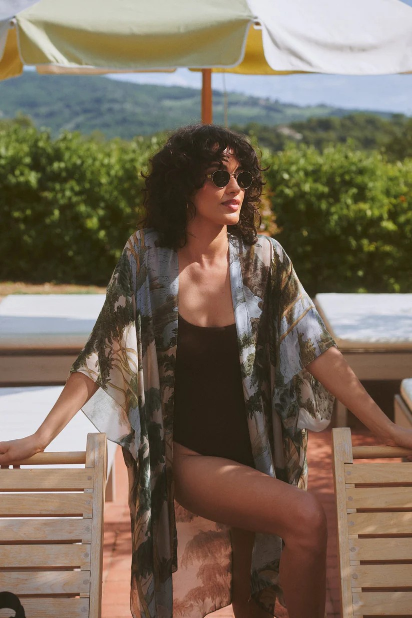 Woman in a floral kimono and sunglasses sitting under an umbrella with a scenic background