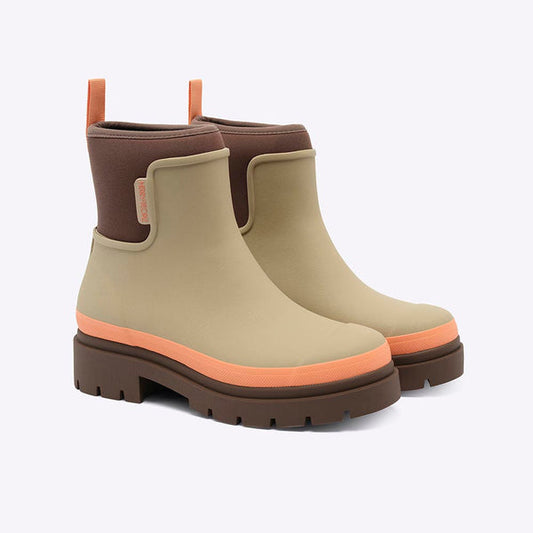 Merry People Tully Ankle Wellies in Taupe & Apricot - a pair of Chelsea-style rain boots in beige, brown, and orange. The boots feature a beige rubber upper, a dark brown elastic side panel, a brown pull tab, and a chunky dark brown lug sole with a bright orange stripe accent. 