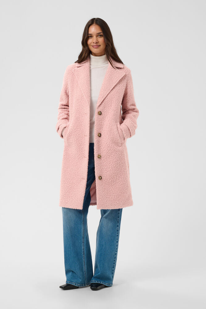 Kaffe Anne Coat in Pink Mauve - a woman modelling a light pink teddy coat with brown buttons, worn over a white turtleneck and blue jeans, against a plain light grey studio background.