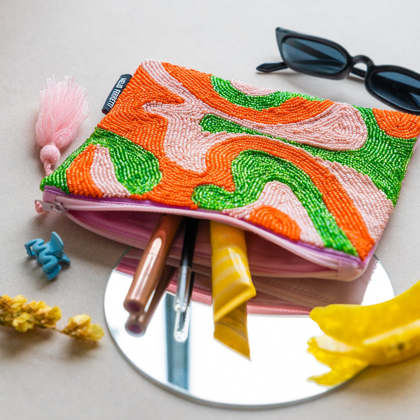 Colorful beaded pouch with sunglasses, hair clips, and makeup items on a mirror.