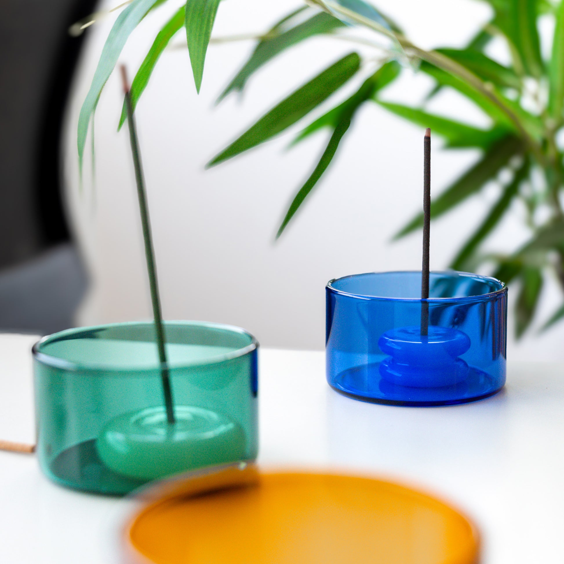Two glass containers, one green and one blue, with incense sticks on a white surface with a blurred plant background.