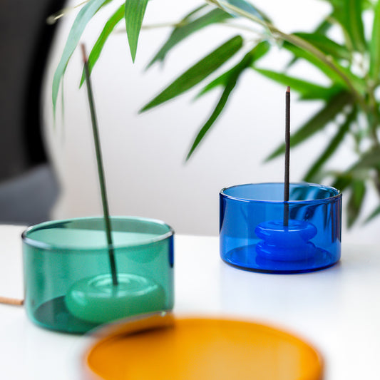 Two glass containers, one green and one blue, with incense sticks on a white surface with a blurred plant background.