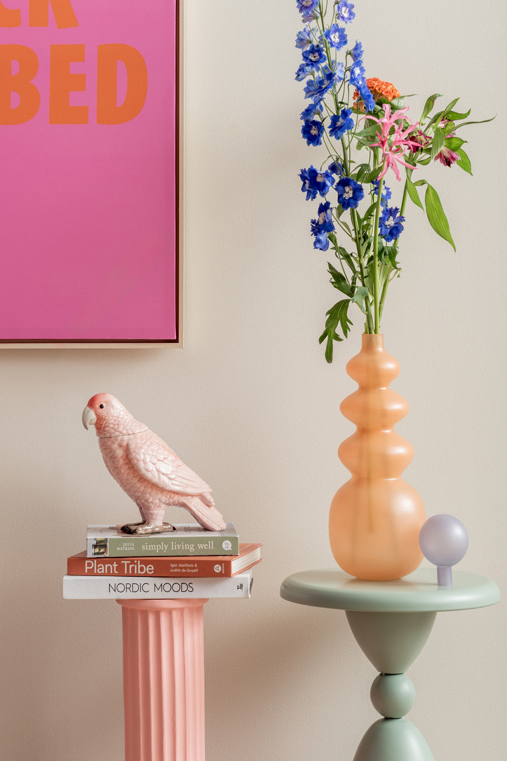 Decorative setup with a parrot figurine, books, and a vase with flowers on a table against a pink wall.
