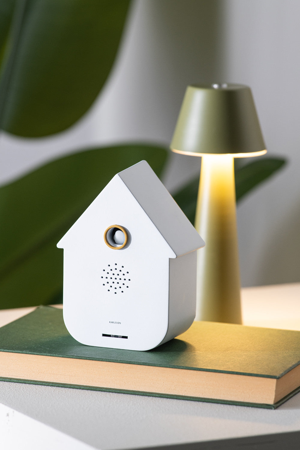 White birdhouse-shaped device on a book with a lamp in the background