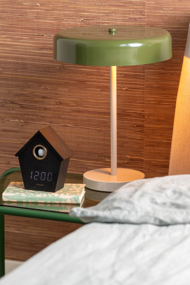 Green table lamp and black clock on a wooden surface with a bed in the foreground