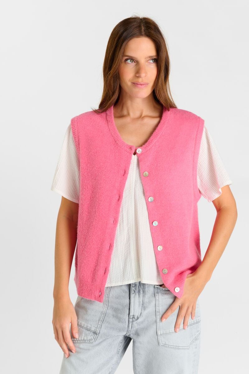 Woman wearing a pink cardigan over a white shirt and light blue jeans on a plain background