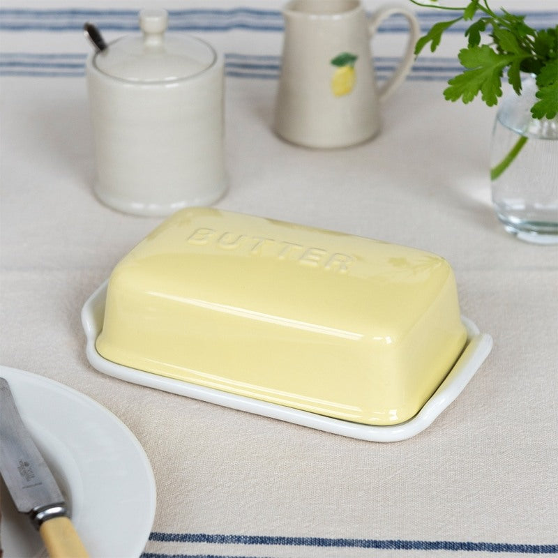 Rex London Ceramic Butter Dish - Yellow - a pale yellow butter dish with a matching butter top, embossed with the word 'BUTTER,' set on a cream tablecloth with blue stripes. A white plate with a knife, a white jar, a white pitcher, and a glass vase with greenery are visible in the background.