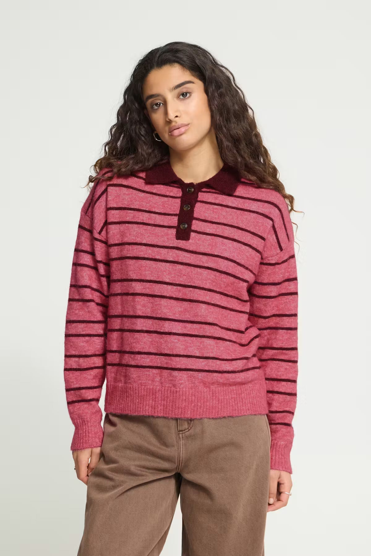 ICHI Sage LS Pullover in Fruit Dove With Zinfandel Stripe - a woman modelling a pink and dark red striped knit polo sweater with a dark red collar, paired with brown pants, against a plain light grey background.