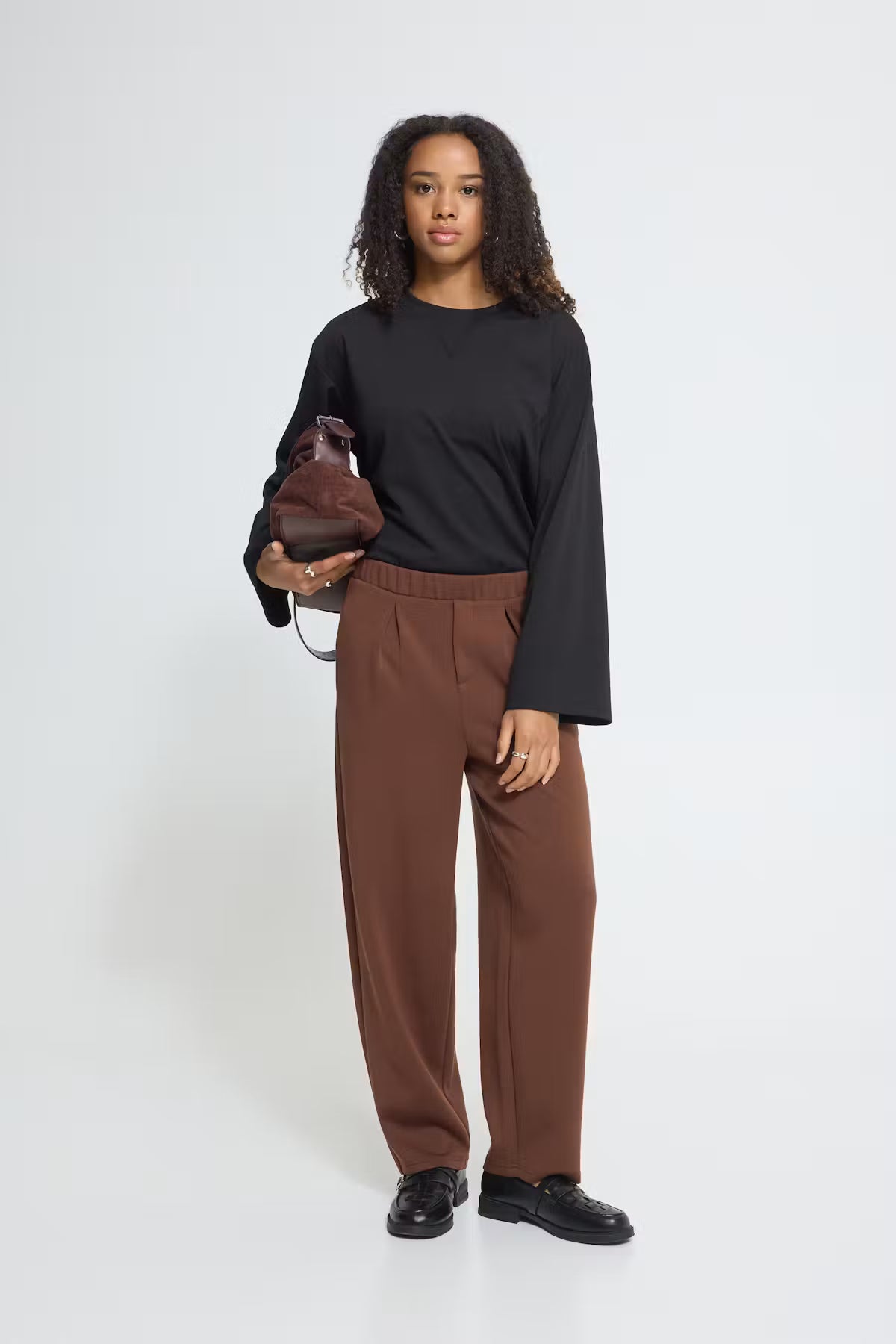 A model stands against a light gray studio background wearing a black long-sleeve top and brown wide-leg trousers, holding a small brown leather bag.
