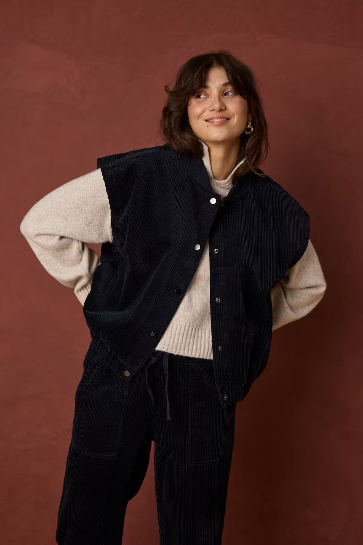 A woman posing in a dark navy corduroy button-up vest over a beige ribbed turtleneck sweater, paired with matching dark corduroy pants, set against a textured reddish-brown studio backdrop.