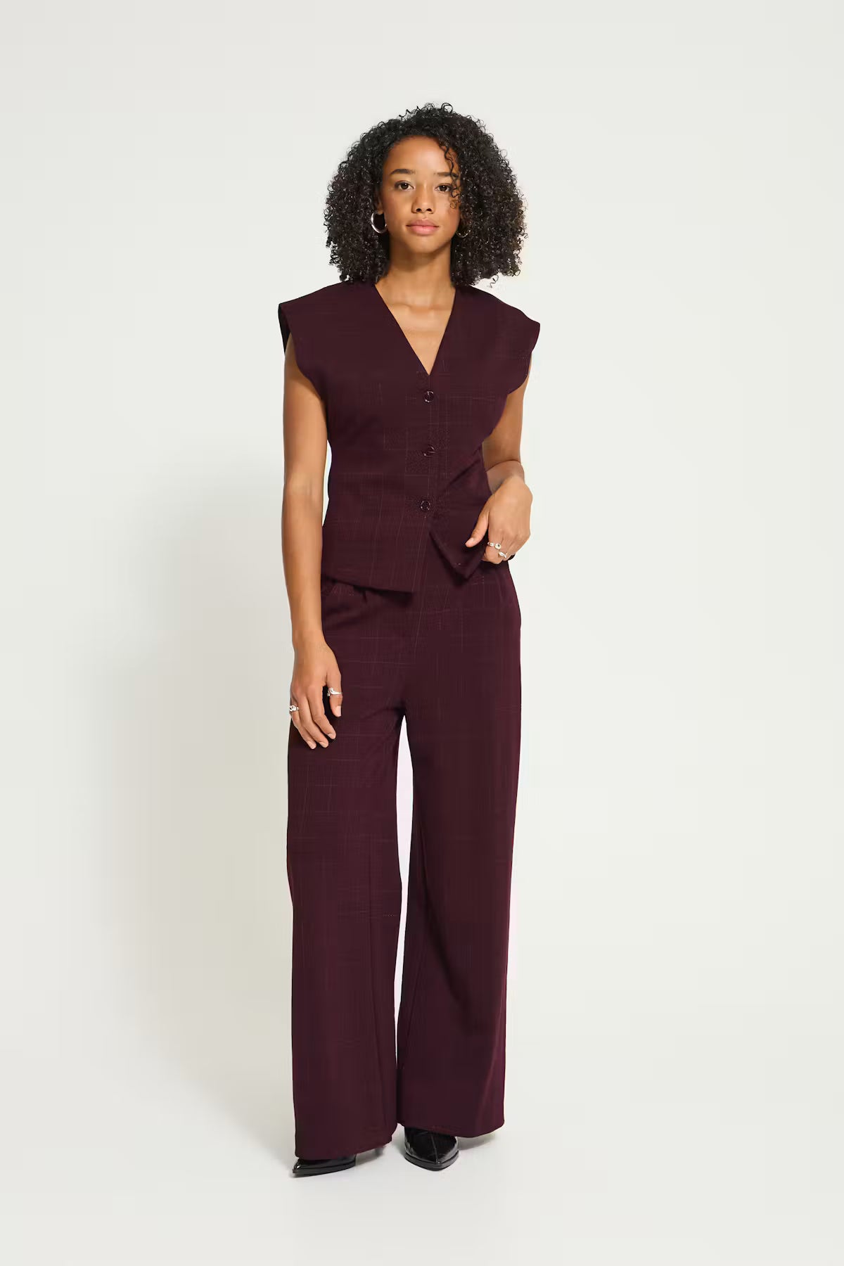A woman modeling a sleeveless dark plum jumpsuit with a V-neck, button front, and wide-leg pants, photographed against a plain light gray studio background.