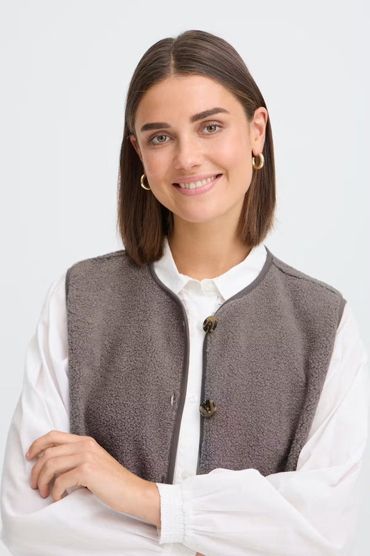 A woman with a warm smile wearing a brown textured fleece vest over a white collared blouse, set against a clean light gray studio background.