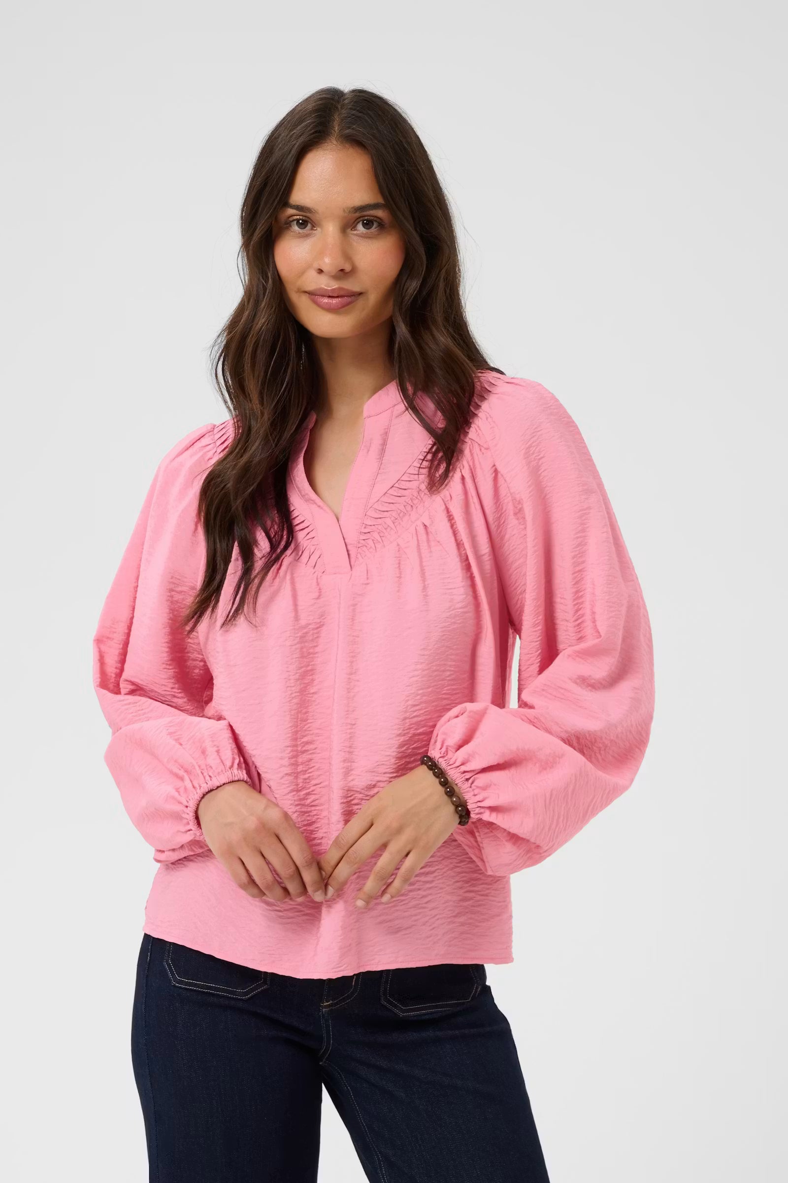 A woman wearing a pink long-sleeve blouse with a gathered, ruffled V-neckline and elasticized cuffs, paired with dark blue jeans, against a light gray studio background.