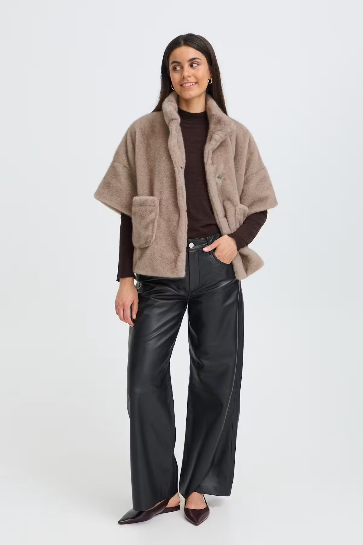 A woman modeling a light brown faux fur cropped jacket over a dark brown top and black wide-leg pants, standing against a light gray studio background.