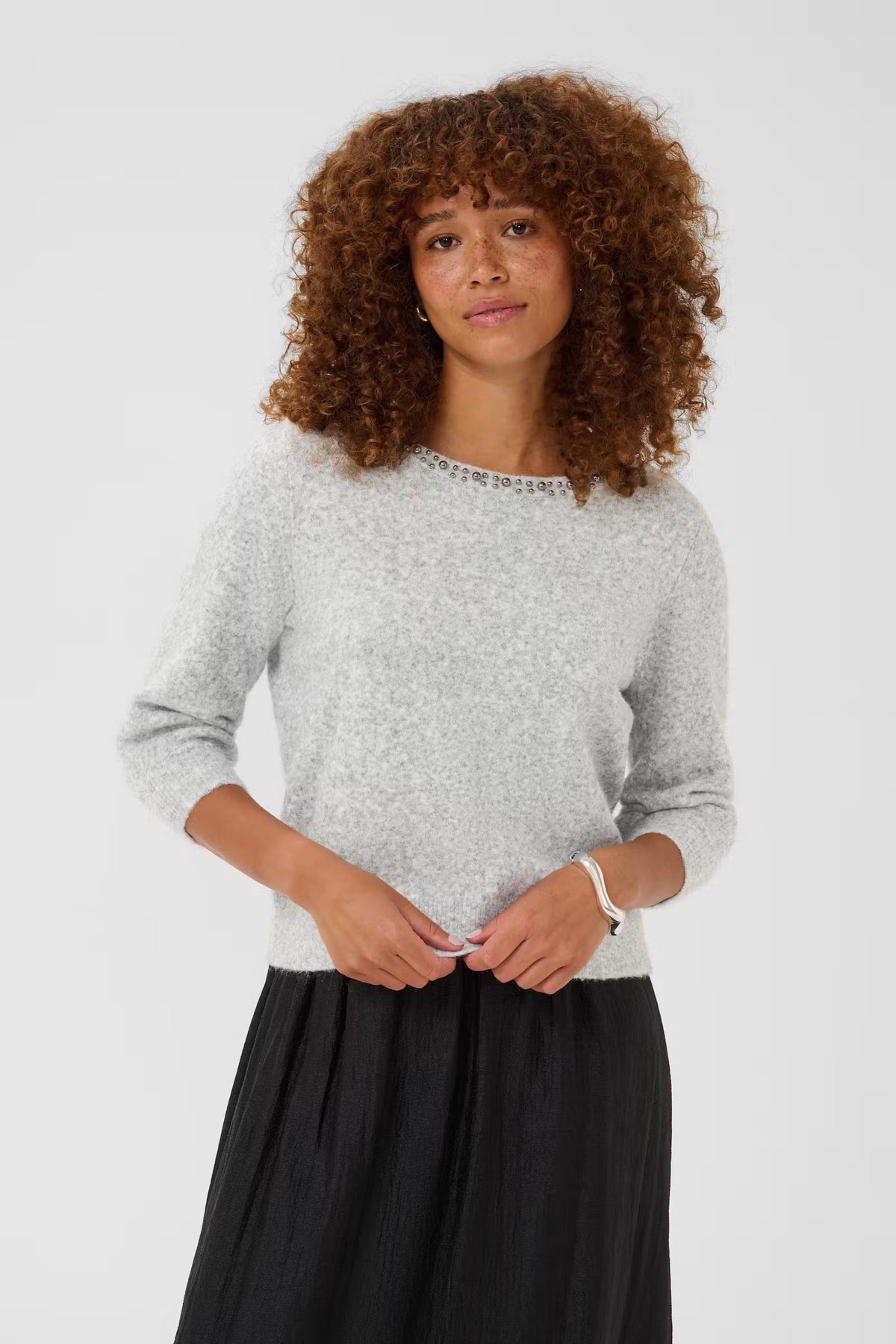 Kaffe Selly Pullover in Grey Melange - A woman with curly brown hair wearing a light grey marled knit sweater with a beaded crew neckline and a black pleated skirt, standing against a light grey studio background.