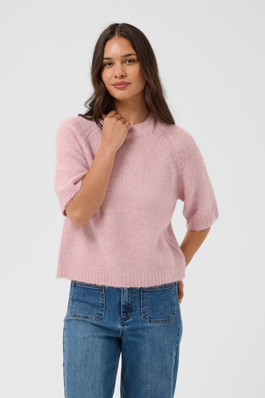 A woman modeling a light pink short-sleeve knit sweater with blue jeans against a plain light gray studio background.