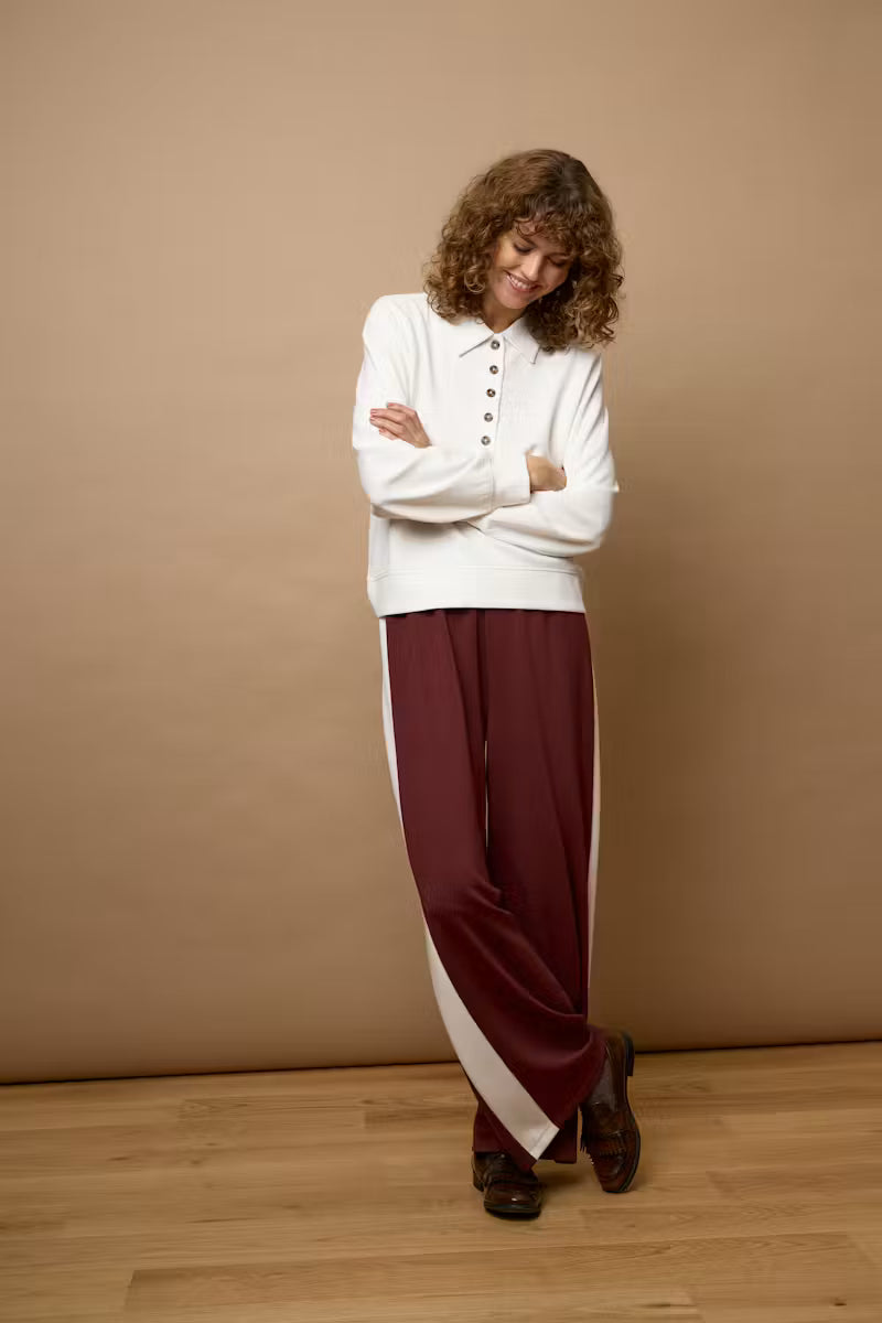 A woman stands with arms crossed wearing a white long-sleeve henley-style top and wide-leg burgundy pants with a white side stripe, posed against a plain beige studio backdrop with a light wood floor.
