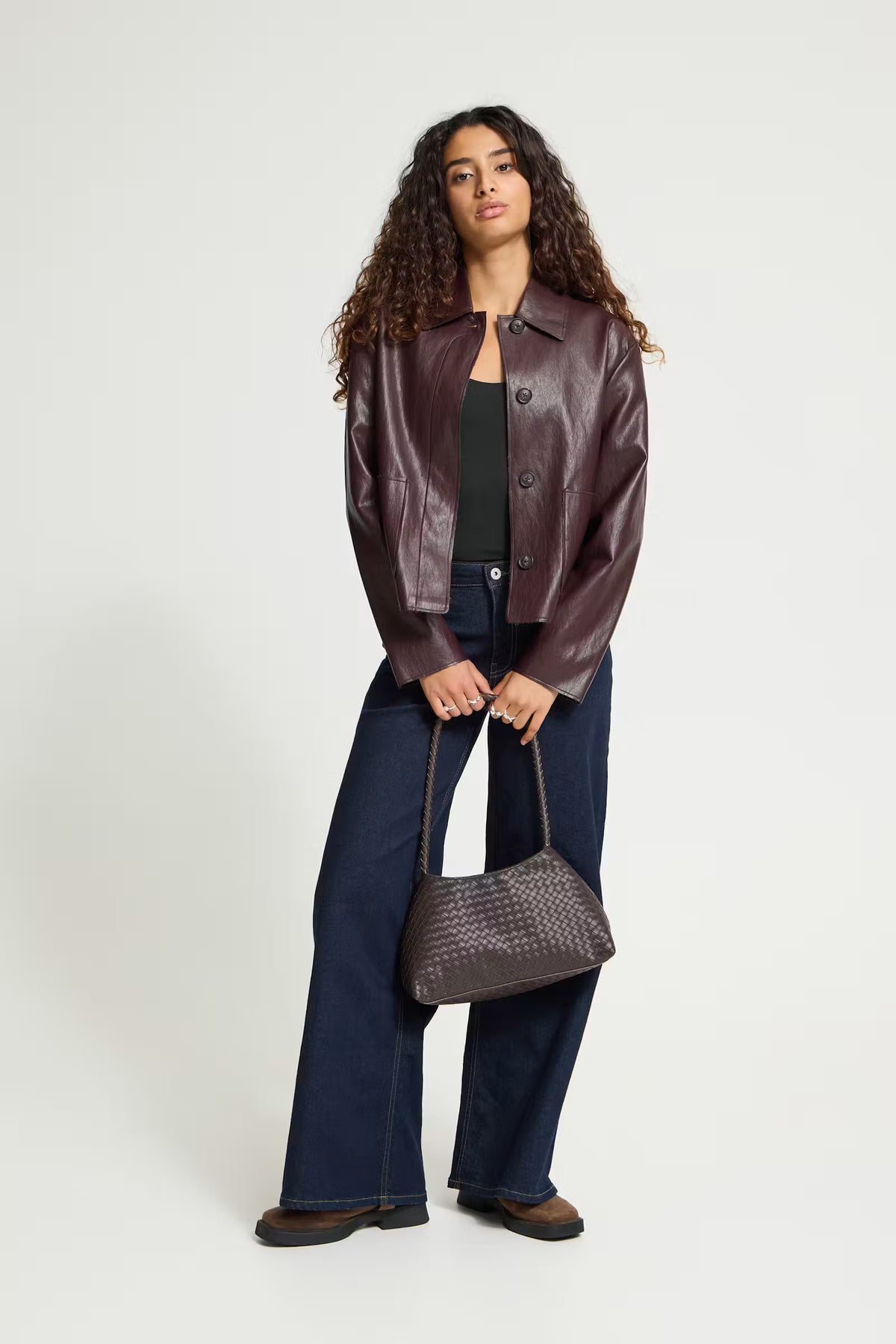 ICHI HARUMI Casual Jacket in Fudge - A model stands against a light grey studio background wearing a dark brown leather jacket, dark blue wide-leg jeans, a black top, and brown shoes, while holding a dark brown woven handbag.