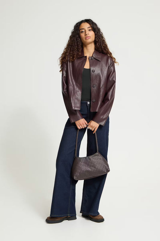 ICHI HARUMI Casual Jacket in Fudge - A model stands against a light grey studio background wearing a dark brown leather jacket, dark blue wide-leg jeans, a black top, and brown shoes, while holding a dark brown woven handbag.