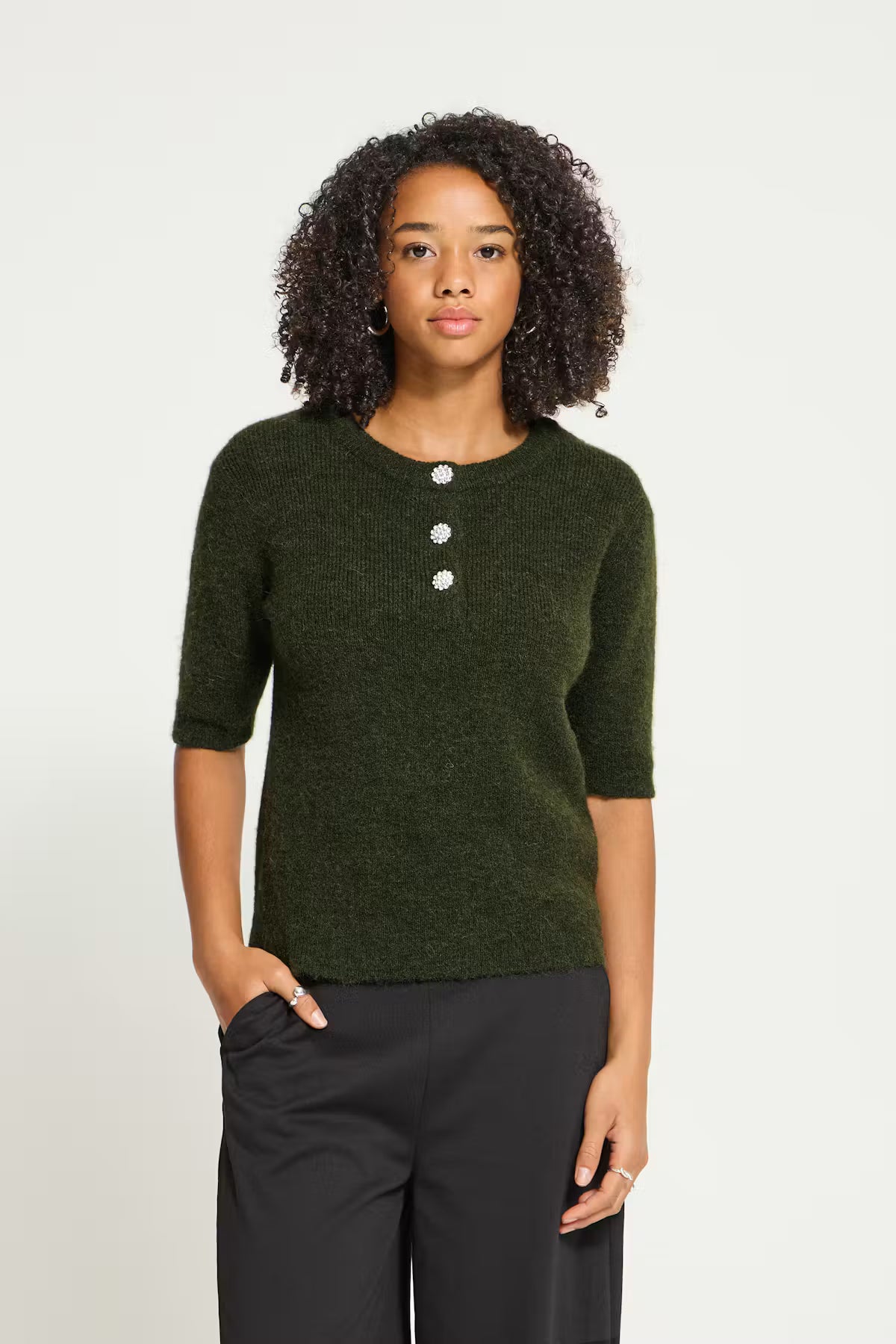 ICHI Fantia Knit in Forest Night Melange - A woman with curly dark hair wearing a dark olive green knit sweater with three white buttons at the henley neckline, paired with black trousers, standing against a light grey studio background.