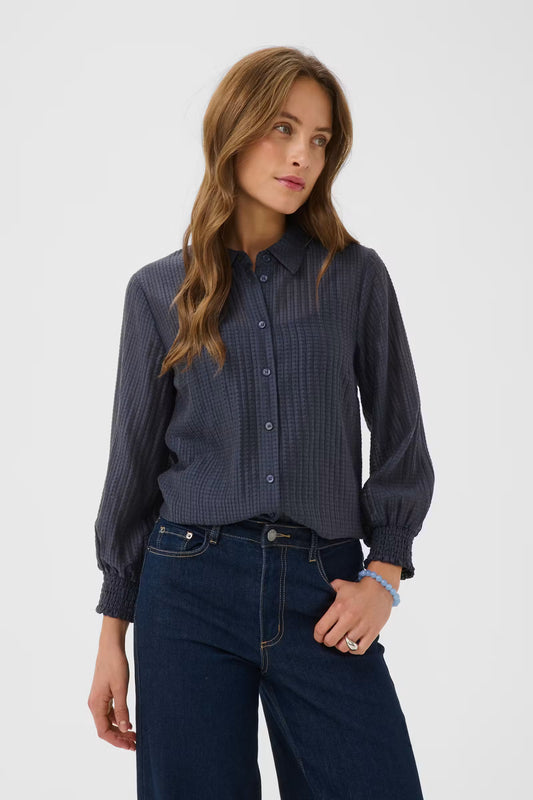 Kaffe Rea Shirt in Graphite - a woman wearing a dark navy blue, textured long-sleeve button-up shirt with dark blue jeans, photographed against a light grey studio background.