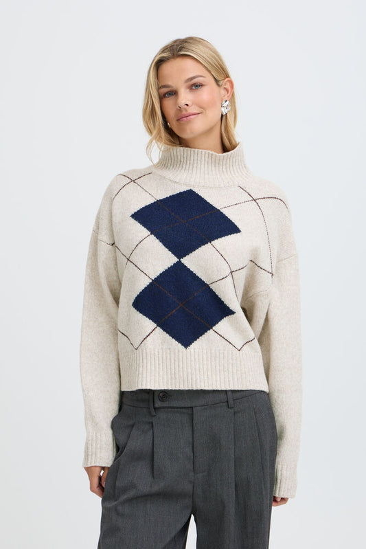 Fransa GIGI Pullover in Oatmeal Melange Check - A woman wearing a cream-colored knit sweater with a navy blue argyle pattern on the front, paired with dark grey trousers, against a light grey studio background.