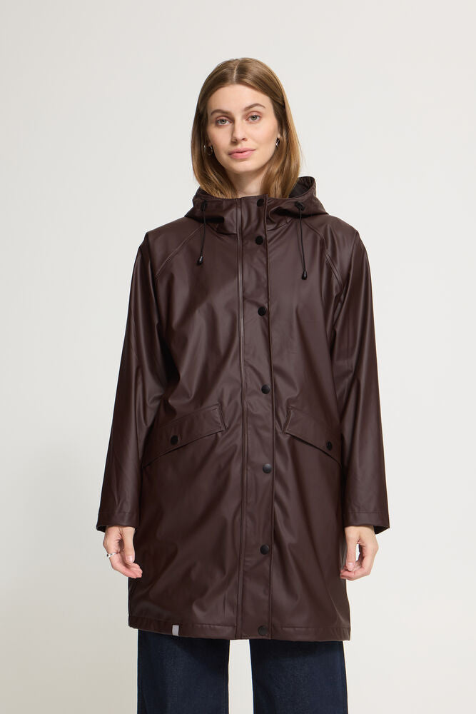A woman wearing a dark brown hooded raincoat with front buttons and flap pockets, paired with dark blue jeans, standing against a light gray studio background.
