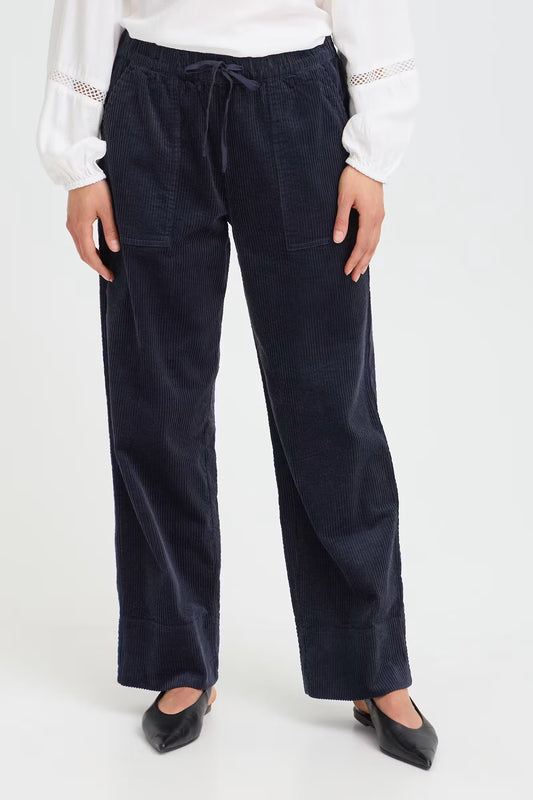 A person wearing dark navy corduroy wide-leg trousers with a drawstring waist, paired with a white blouse and black pointed flats, shown from the waist down against a light gray studio background.