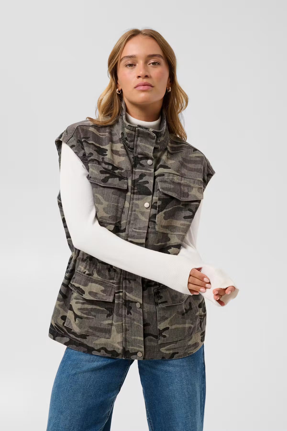 Kaffe Lilana Warm Waistcoat in Camo - a woman wearing a sleeveless camouflage utility vest over a white long-sleeve turtleneck and blue jeans, standing against a light grey studio background.
