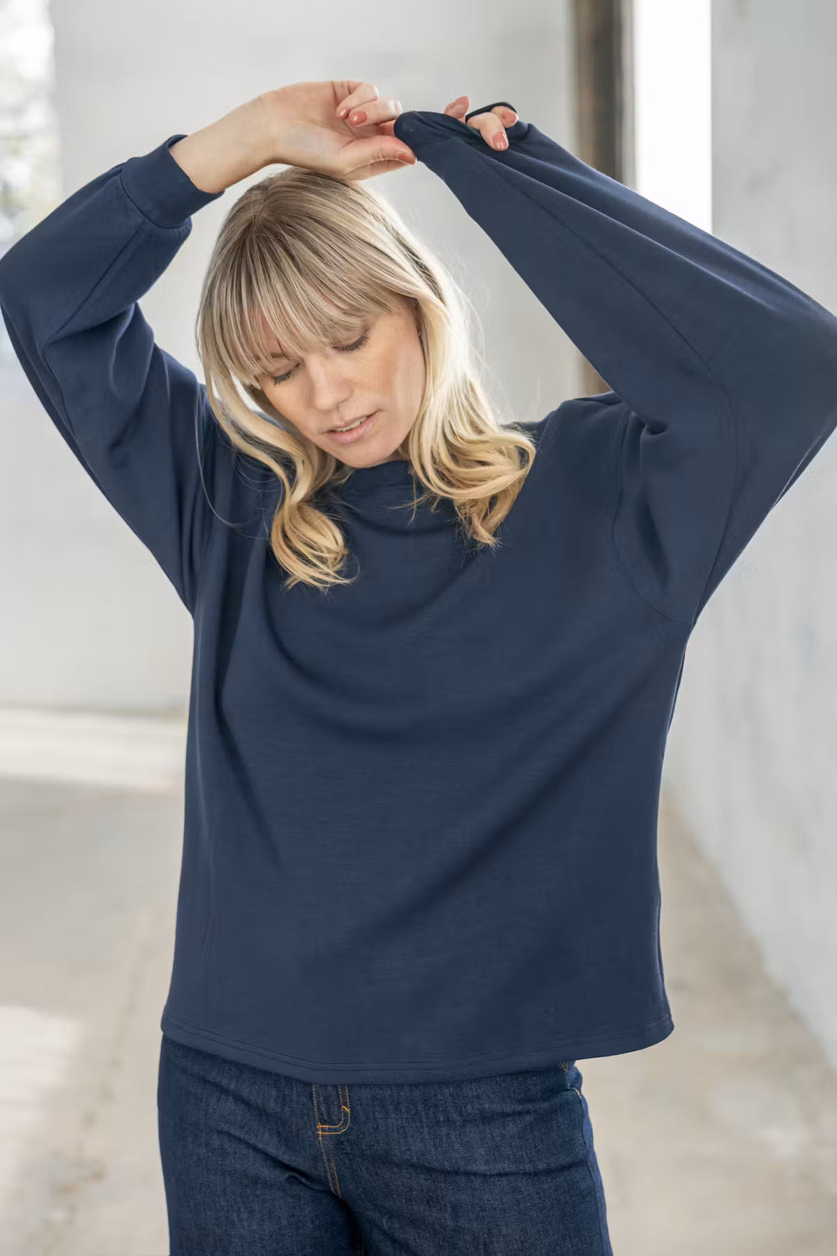 Fransa Aurora Pullover in Navy Blazer - A woman wearing a navy blue long-sleeve mock neck top and dark blue jeans, posing with her hands raised above her head in a bright, modern indoor setting.