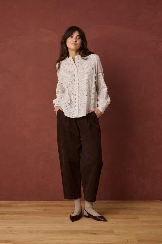 A woman stands against a textured terracotta wall on a light wood floor, wearing a white long-sleeve lace blouse, dark brown cropped trousers, white socks, and dark brown pointed flats.