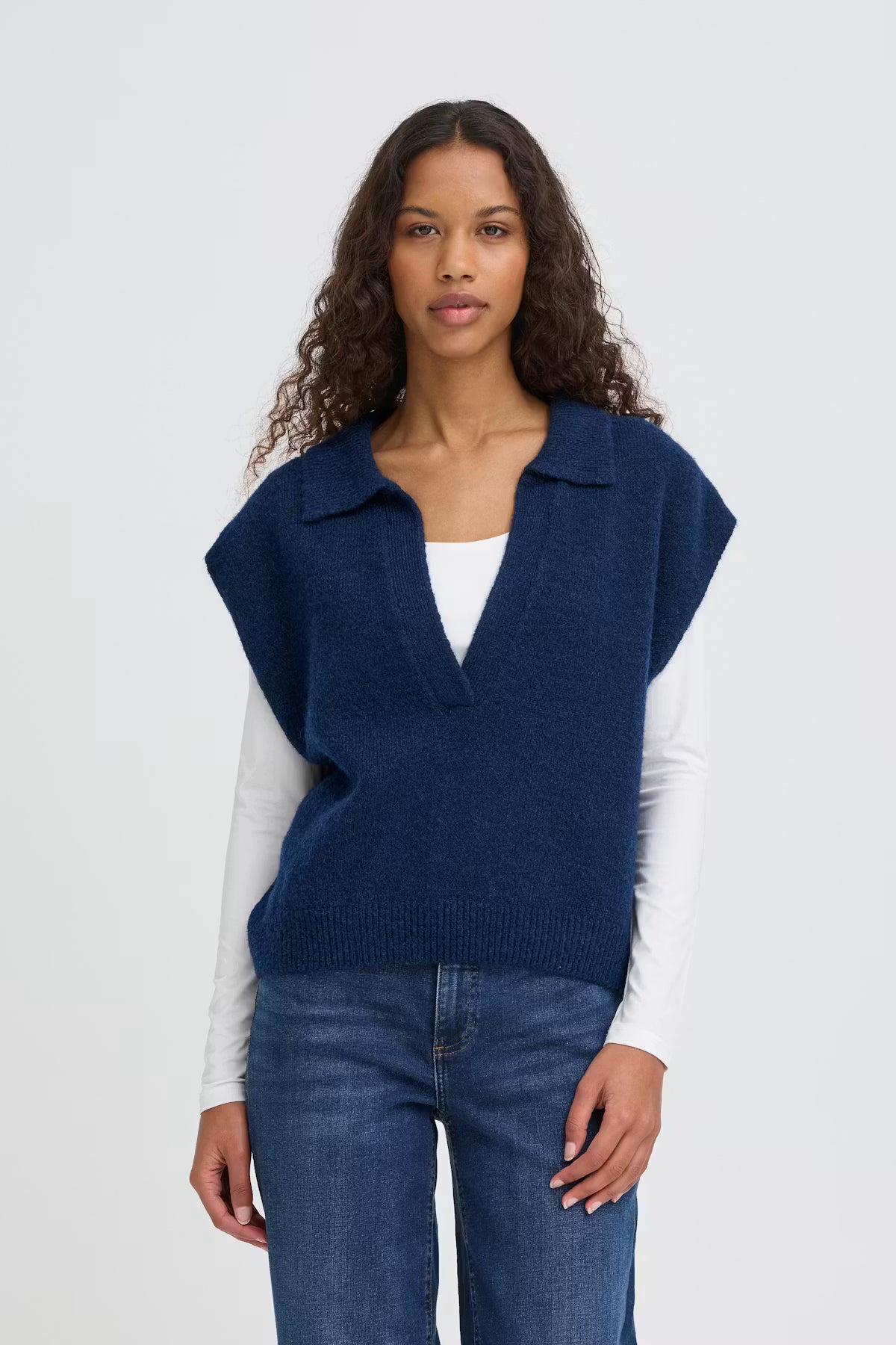 Woman wearing a blue vest over a white shirt and jeans on a light gray background
