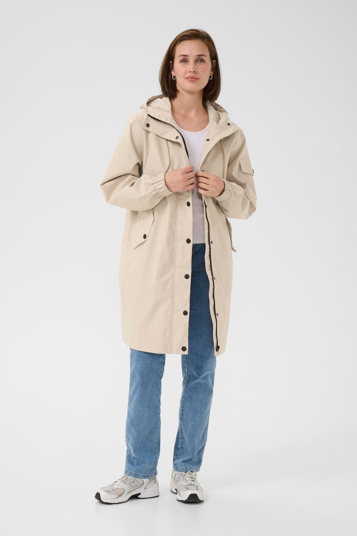 Woman wearing a beige raincoat, white shirt, blue jeans, and gray sneakers on a white background
