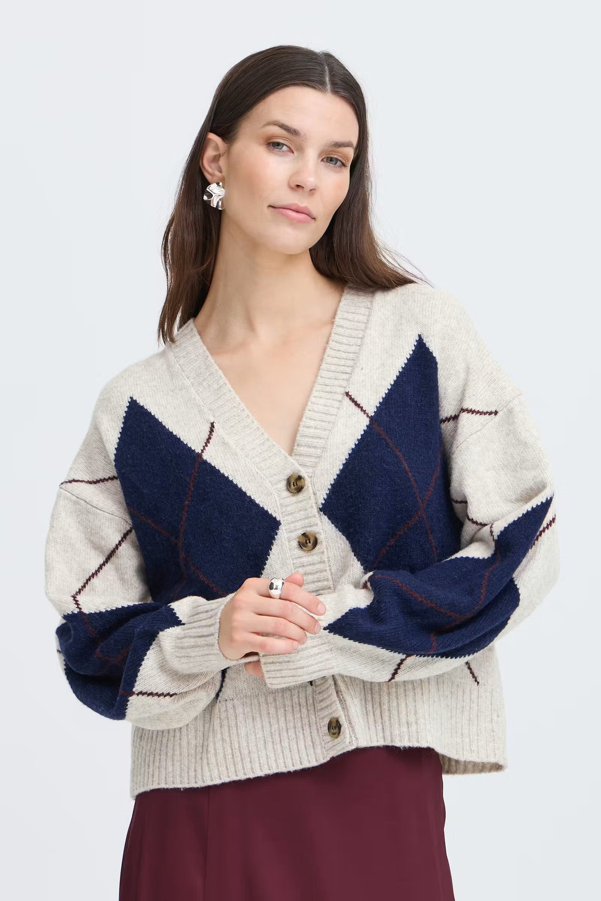 Fransa GIGI Cardigan in Oatmeal Melange Check - A woman wearing a cream and navy blue argyle cardigan sweater with brown buttons, paired with a burgundy skirt, against a light grey studio background.