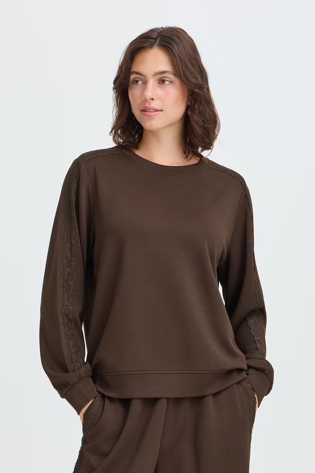 Fransa Aurora Sweatshirt in Mole - A woman wearing a dark brown crewneck sweatshirt with textured lace detailing on the sleeves, paired with matching brown pants, against a light grey studio background.