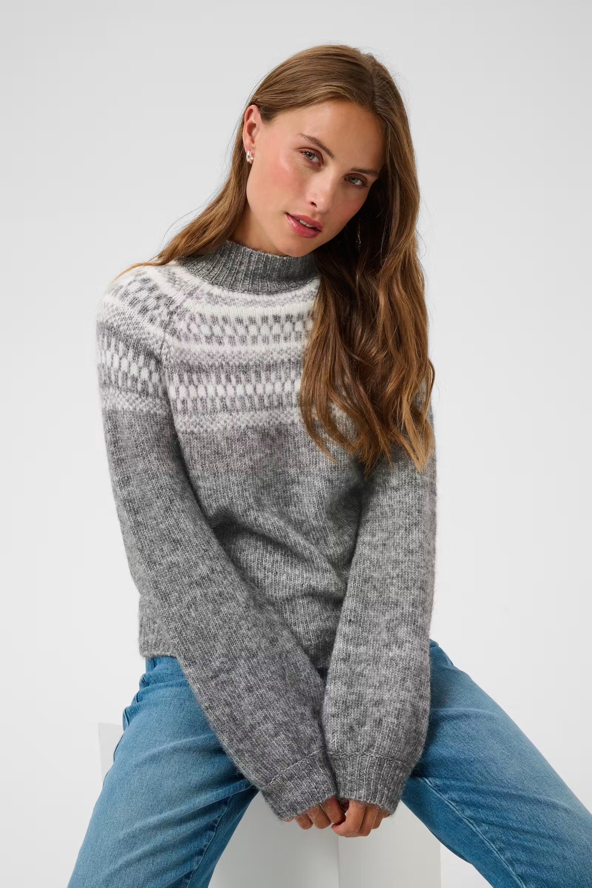 A woman wearing a gray and white patterned knit sweater with blue jeans, seated against a plain light gray studio background.