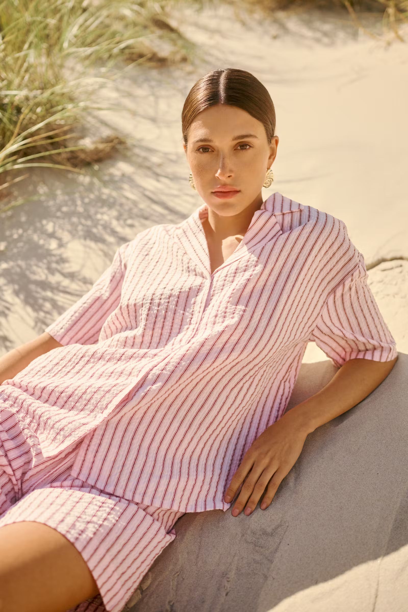 Woman in a pink striped outfit sitting on sand with grass in the background
