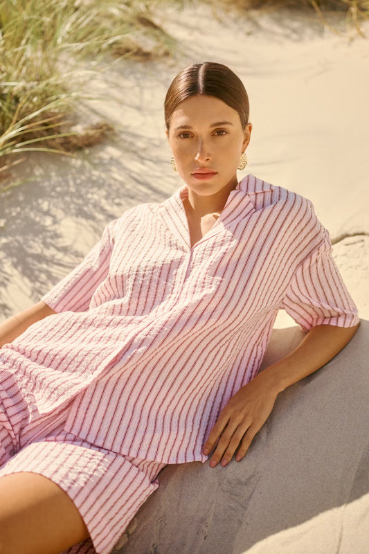 Woman in a pink striped outfit sitting on sand with grass in the background